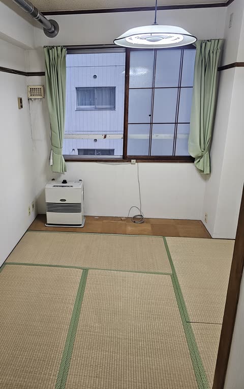 An unfurnished room with tatami mat flooring, a window with green curtains, and a portable kerosene heater in the corner.