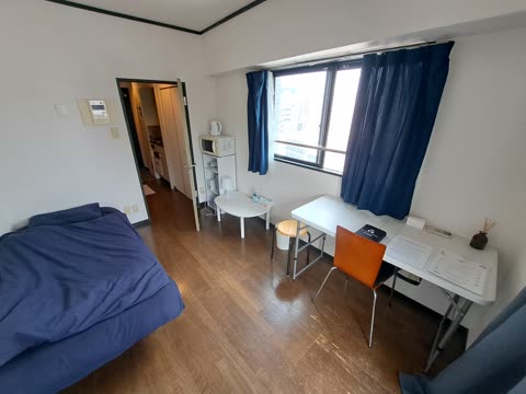 A small, furnished room with wood floors contains a bed with a blue comforter, a desk by a window, and an open doorway.