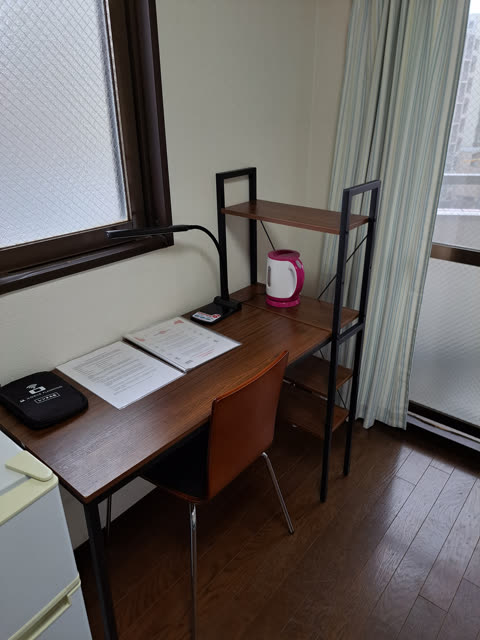A wooden desk with a chair, lamp, and kettle stands in a room next to a window with striped curtains.