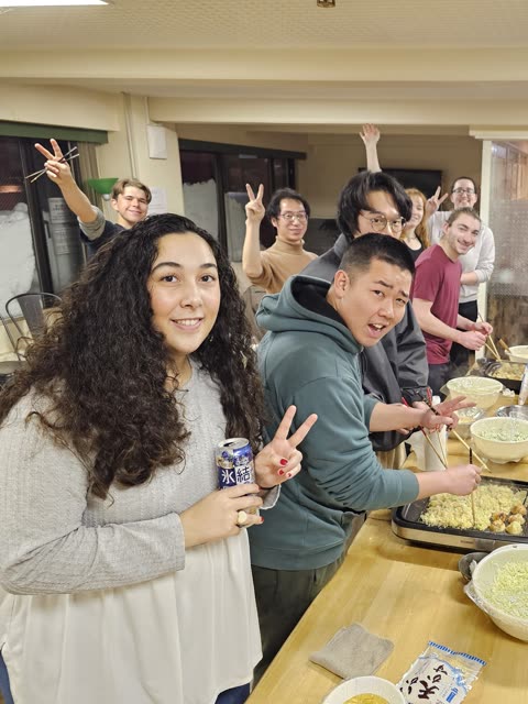 A group of people smile and make peace signs while gathered around a table cooking food on an electric griddle.