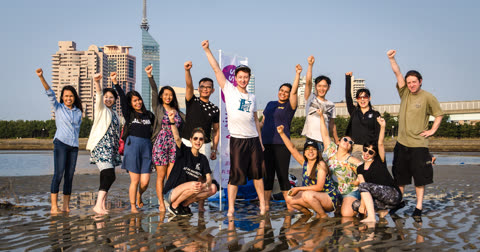 A group of people pose with their fists in the air on a wet, sandy beach in front of a city skyline.