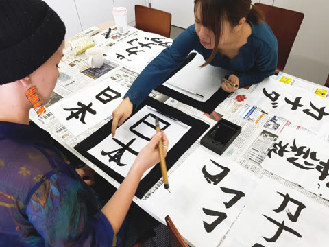 A woman in a blue shirt points to a Japanese character on paper for another person holding a calligraphy brush at a table.