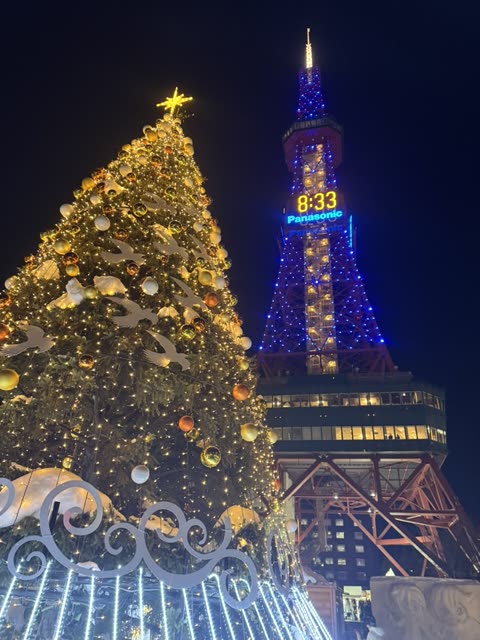At night, an illuminated Christmas tree with gold ornaments stands beside a blue-lit lattice tower displaying the time.