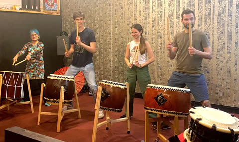 Four people stand holding large drumsticks in front of individual taiko drums in a practice room.