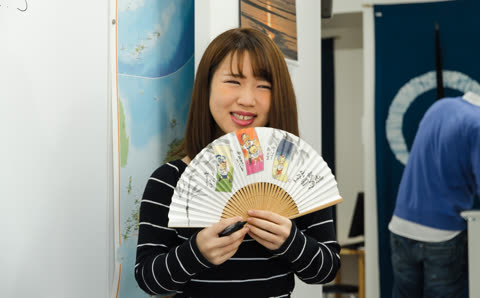 A woman in a striped shirt smiles as she holds up an illustrated paper fan and a marker indoors.