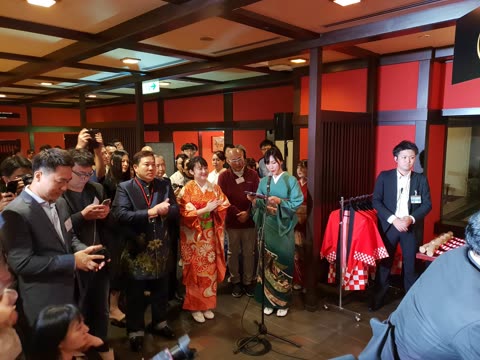 Woman in kimono reading from a tablet at a microphone in a restaurant.