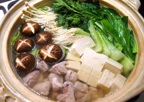 Pot of simmering broth containing tofu, chicken, shiitake mushrooms, and various greens.