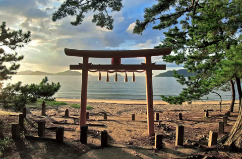 A red torii gate stands on a sandy beach, facing a body of water with distant mountains visible under a cloudy sky.