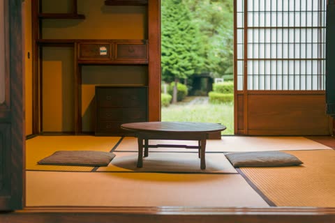 A low wooden table and two floor cushions sit on tatami mats in a room with an open shoji door showing a green garden.