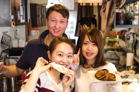 Three people smiling in a kitchen; two women hold food.