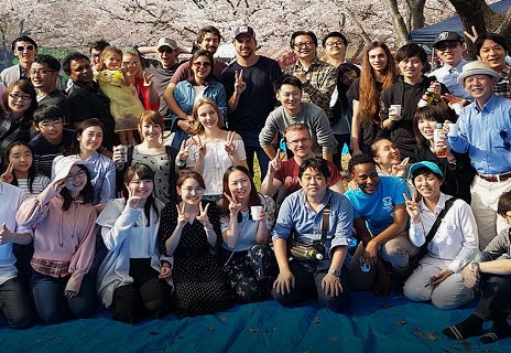 A large group of people poses for a group photo on a blue tarp outdoors under blossoming cherry trees.
