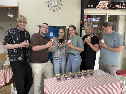 A group of six people stands behind a striped table in a shop, each holding a small, decorative food replica.
