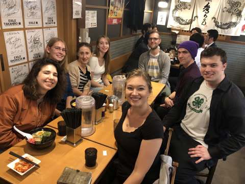 A group of eight people smiles for the camera while seated at a long wooden table inside a Japanese restaurant.