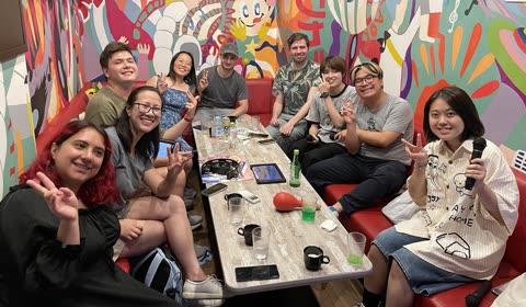 A group of ten people smiles and makes peace signs while sitting around a table in a karaoke room with a colorful mural.