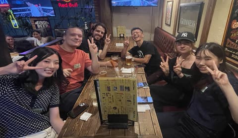 Six people sit at a wooden table in a pub, smiling and making peace signs towards the camera.