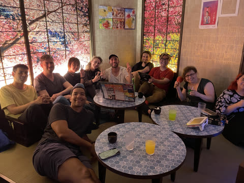 A group of ten people poses for a photo while seated around small tables in a room with cherry blossom wall panels.