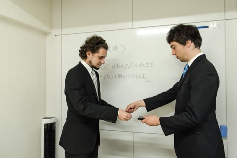 Two men in business suits exchange cards in front of a whiteboard with Japanese characters written on it.