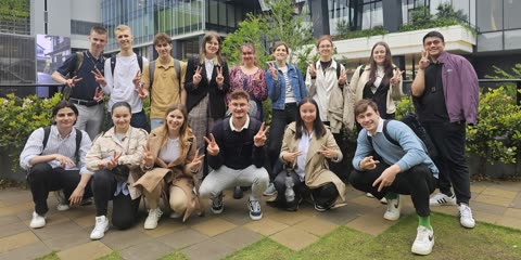 Fourteen people, some standing and some crouching, make peace signs while posing outdoors in front of a glass building.