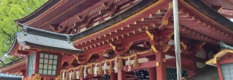 A red, multi-tiered roof of a Japanese-style building with gold accents and decorative ropes is set against green trees.