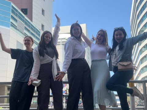 Five young women smile and pose with raised arms on an outdoor urban walkway between tall buildings.
