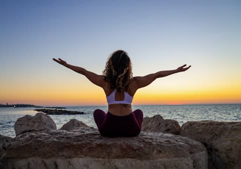 Woman sits on rocks by ocean, arms outstretched at sunset.