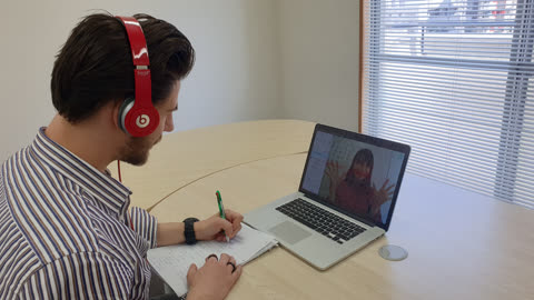 Person wearing headphones writes notes while on a video call at a desk.