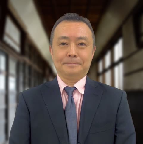 A man in a dark suit, pink shirt, and a tie stands and looks at the camera inside a long, windowed hallway.