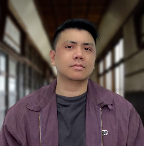 A man in a purple jacket looks at the camera while standing in a long, windowed hallway.