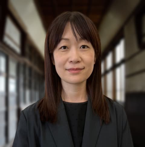 An East Asian woman with brown hair and a dark blazer looks toward the camera in a long, blurred hallway.