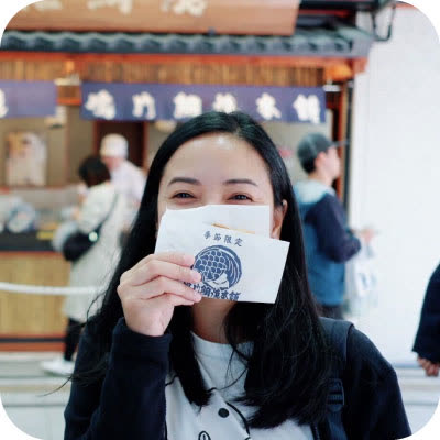 A person with long dark hair holds a small paper bag with a blue logo over their mouth in front of a Japanese storefront.
