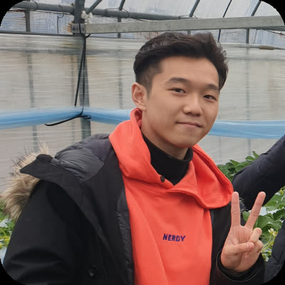 A young man in an orange hoodie makes a peace sign while standing inside a greenhouse.