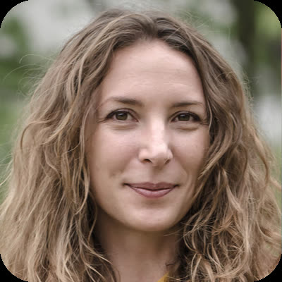 A woman with long, wavy brown hair smiles slightly while looking at the camera in an outdoor setting.