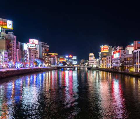 Brightly lit buildings of Fukuoka at night with colorful lights from signs and windows reflecting on a wide river.